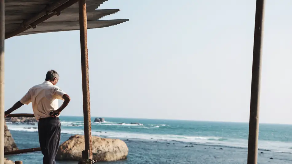 Man leaning on the side railing of a basic roofed shelter and looking out to the sea